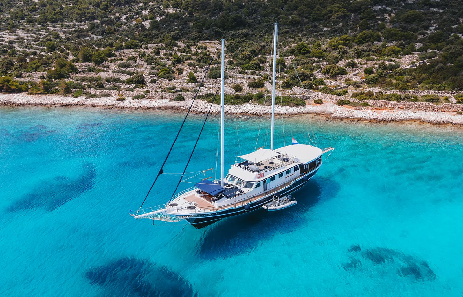Large sailboat anchored in turquoise coastal waters near a rocky, scrub-covered shore.