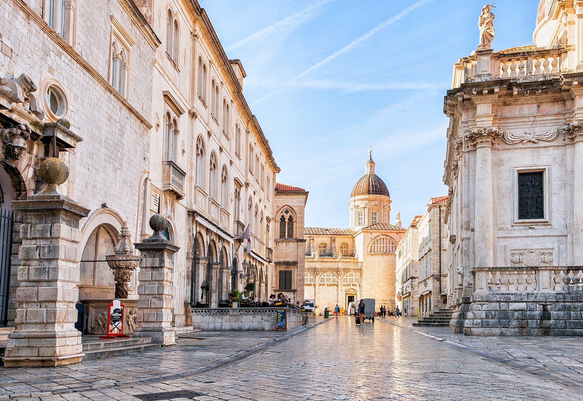 Cobblestone street in Dubrovnik leading to a cathedral with a dome under a blue sky.