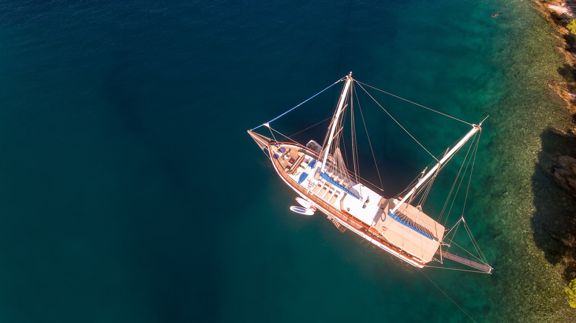 Aerial view of a wooden gulet boat anchored near a rocky, clear-water coastline.