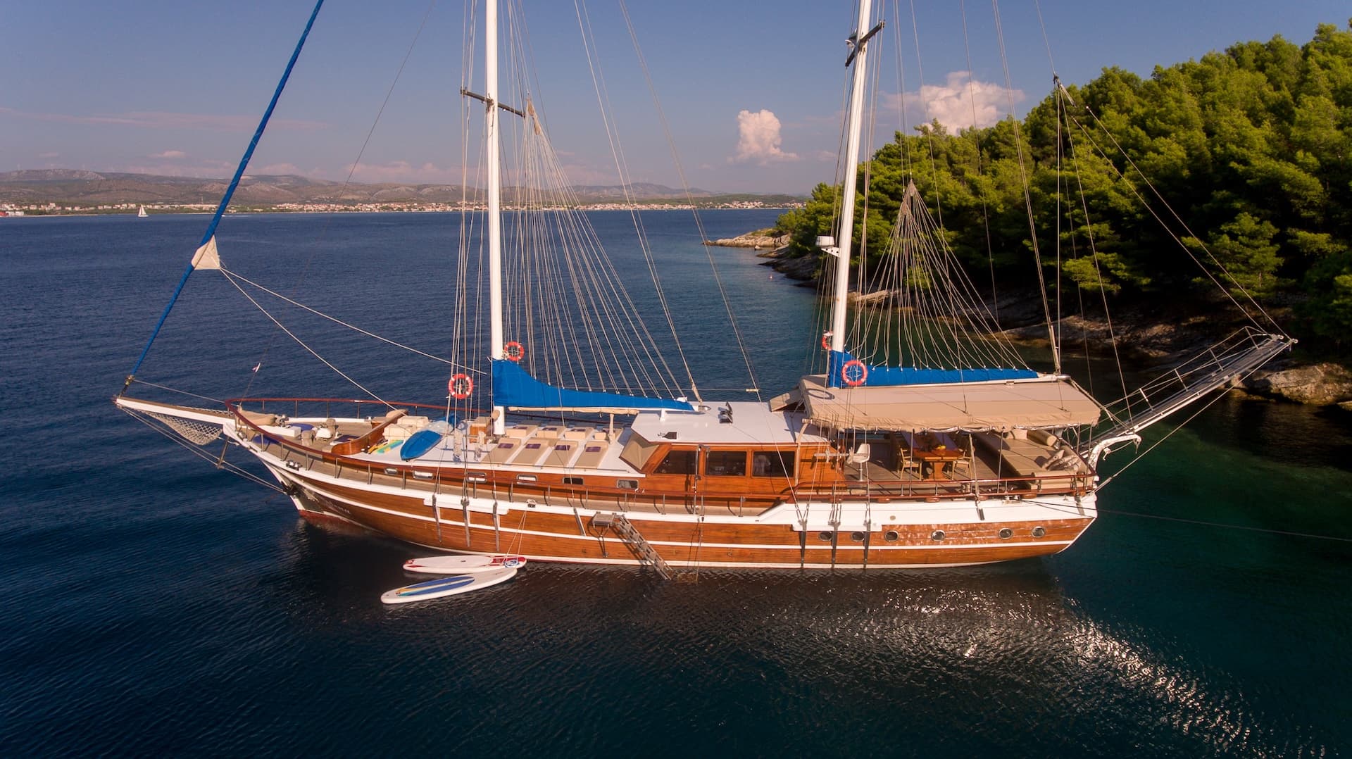 Wooden gulet sailboat anchored near a forested coastline with distant town visible.