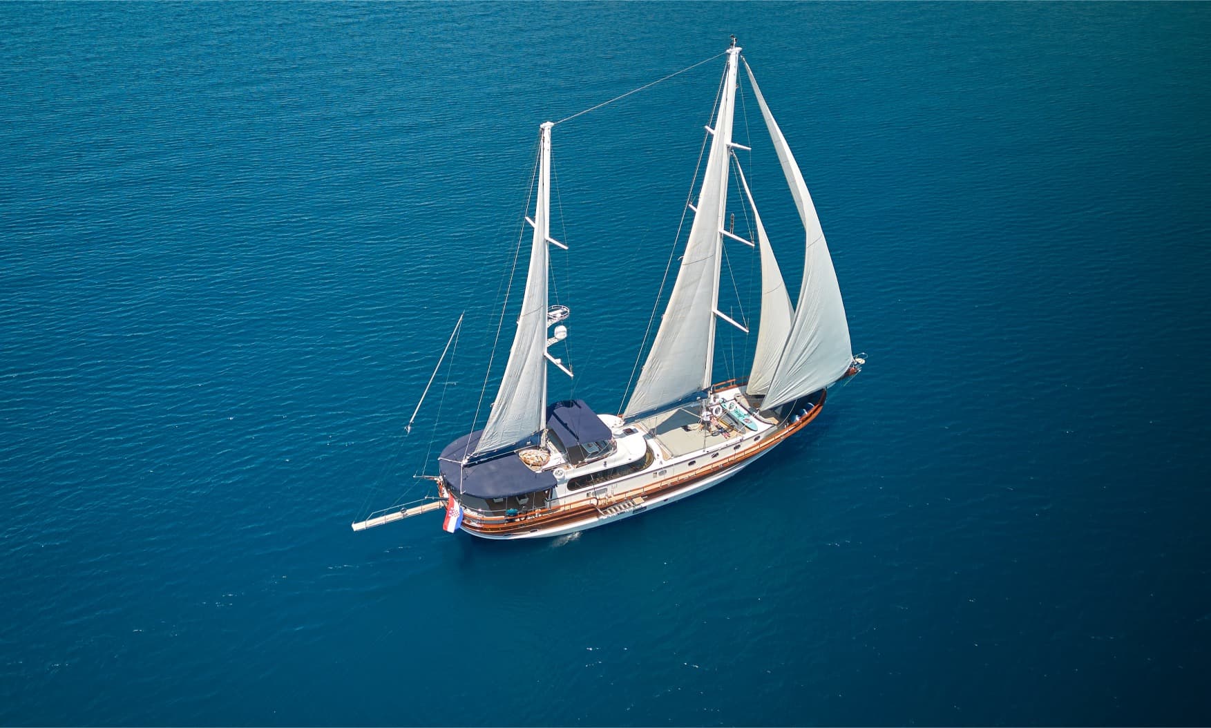 Two-masted wooden sailboat with white sails on deep blue, rippling sea water.
