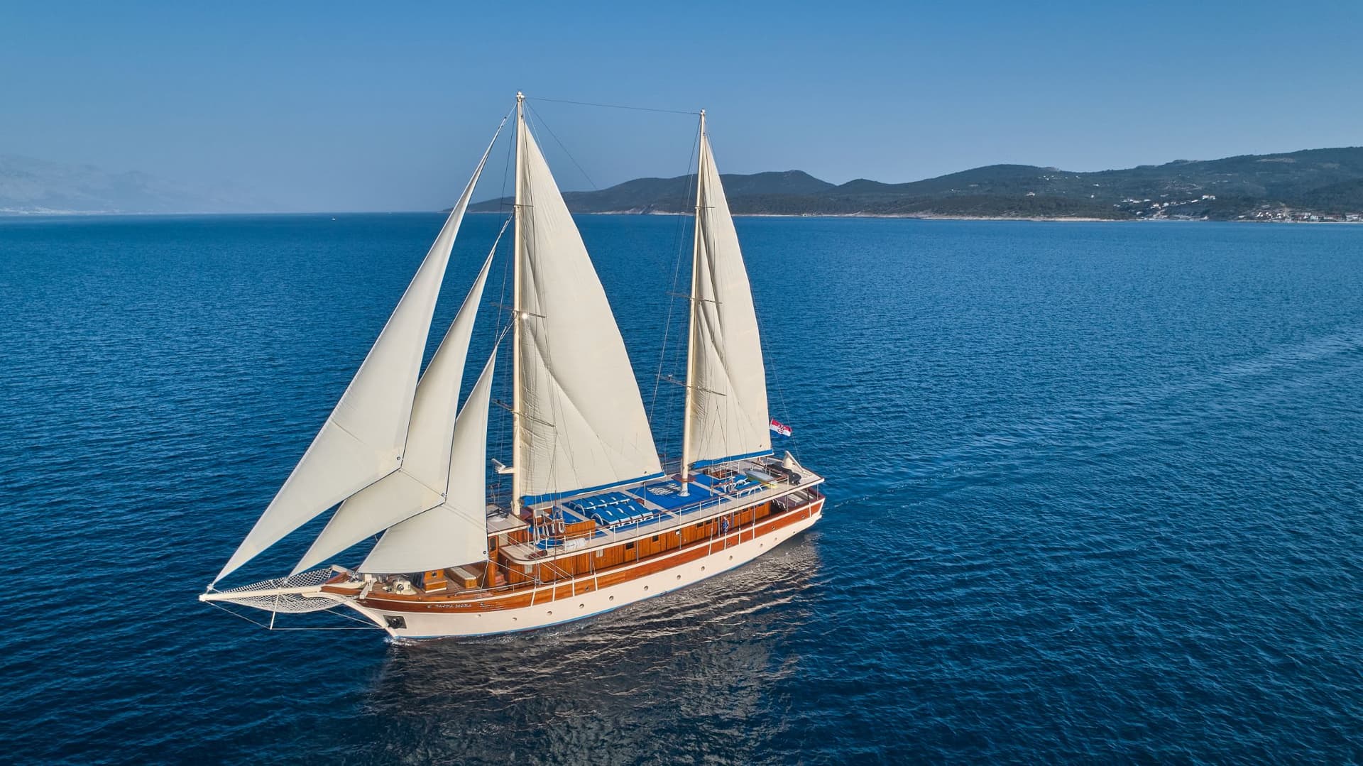 Large wooden sailboat with white sails cruising on deep blue sea near hilly coastline