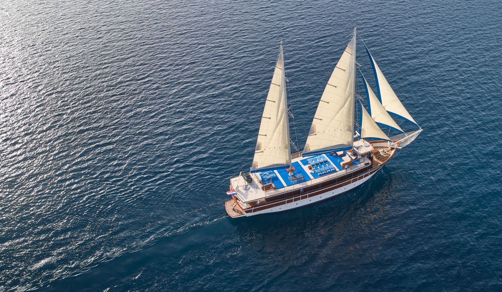 Large wooden sailboat with white and blue sails on deep blue, sunlit sea water.