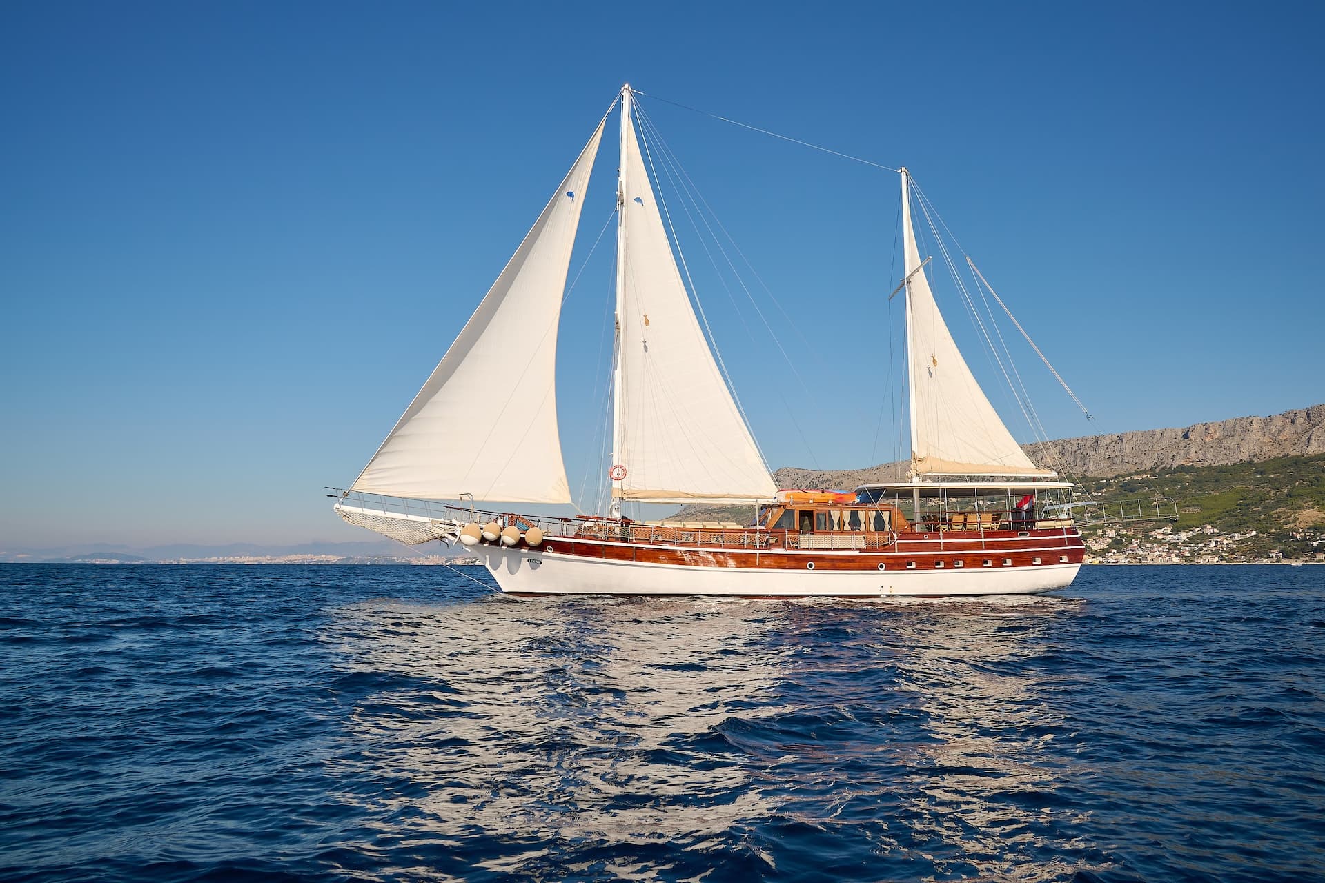 Wooden sailboat with white sails on deep blue water near a rocky coastline.