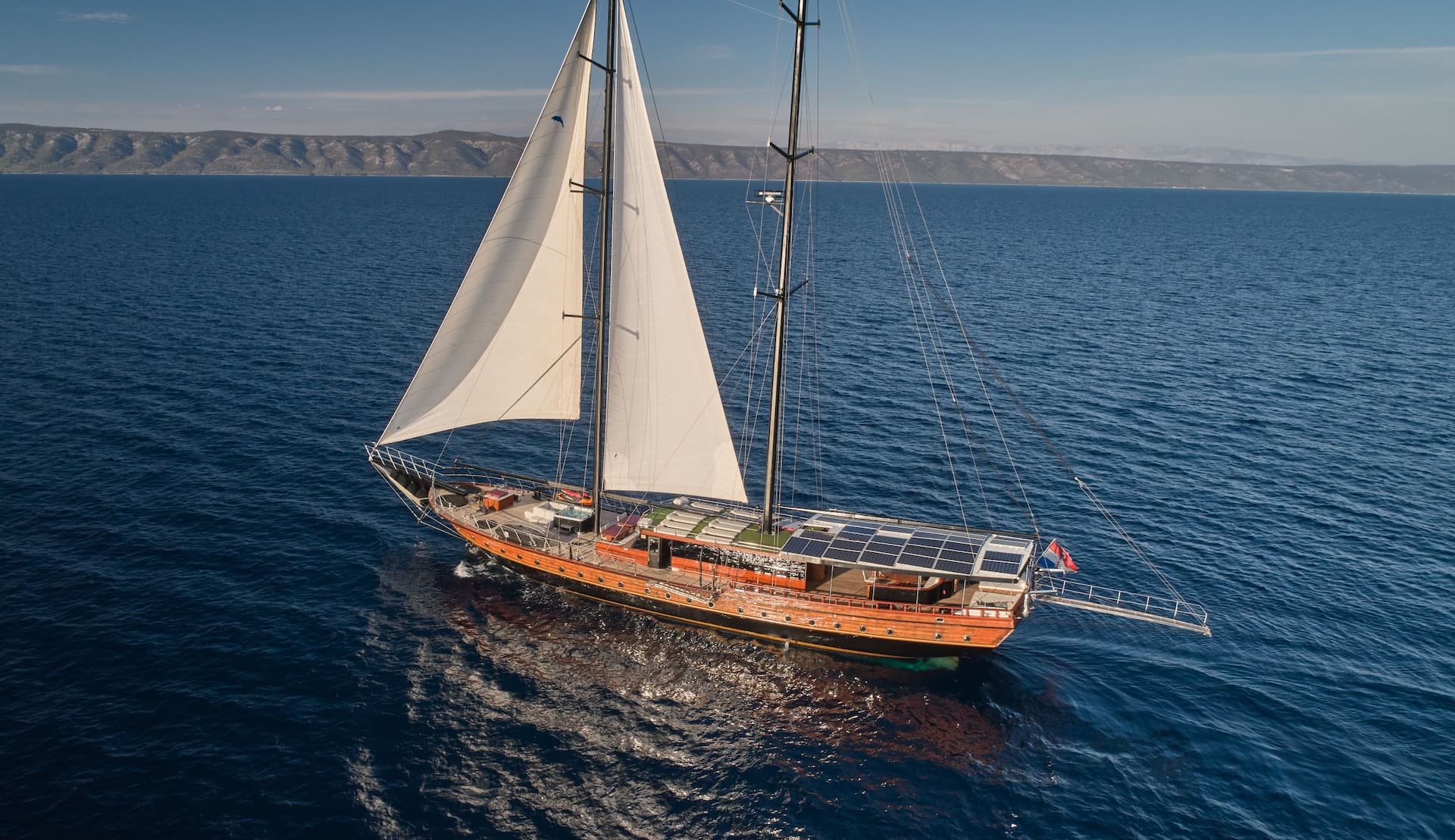 Wooden sailboat with white sails cruising on deep blue sea near a distant coastline.