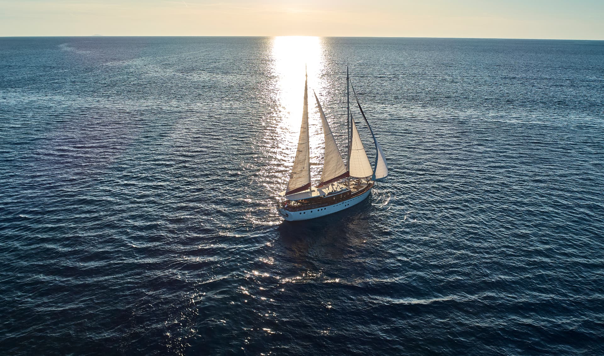 Sailboat with white sails on dark blue sea water during sunset with sun glare.