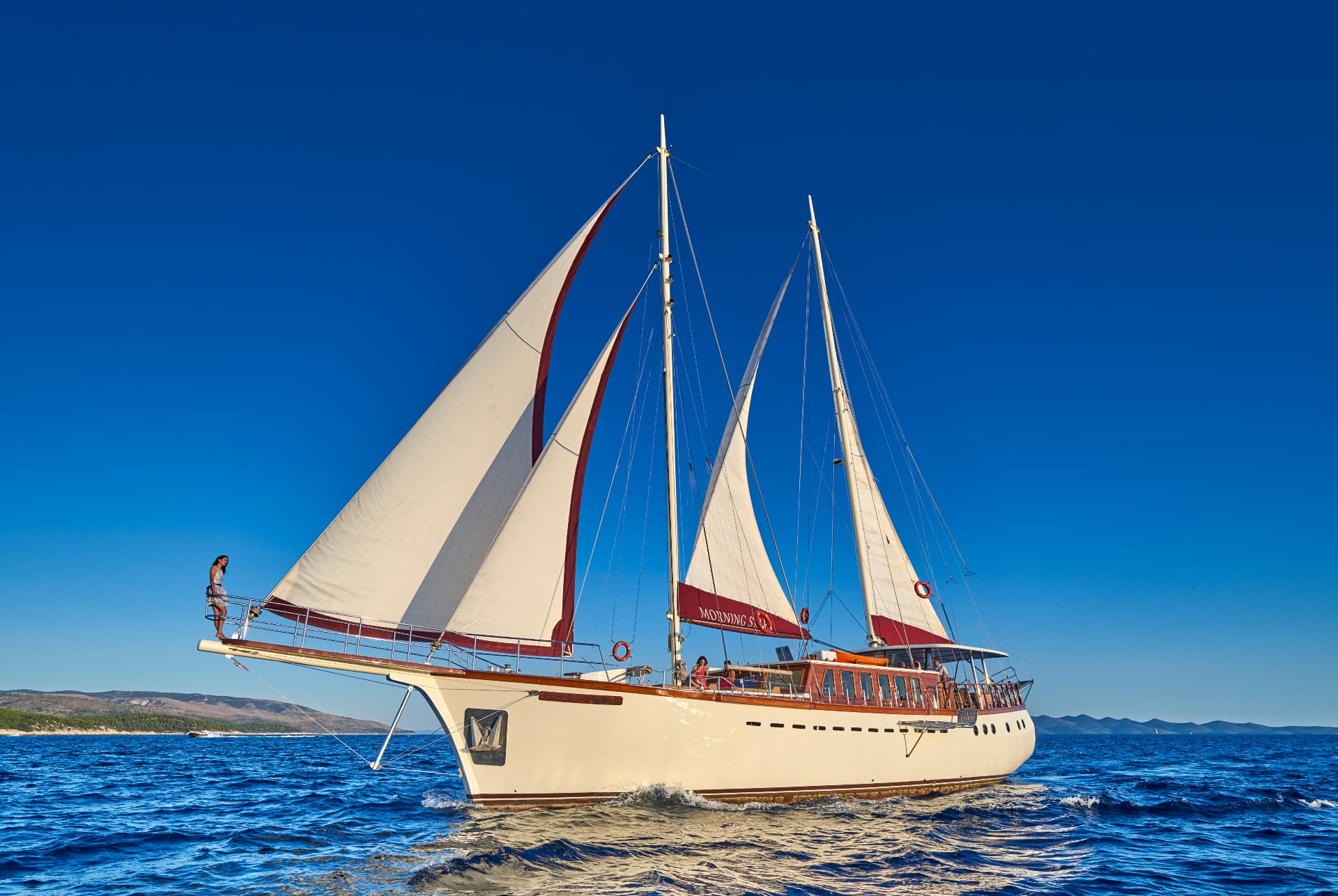 Large white sailboat named Morning Star sailing on deep blue choppy sea near hilly coastline.