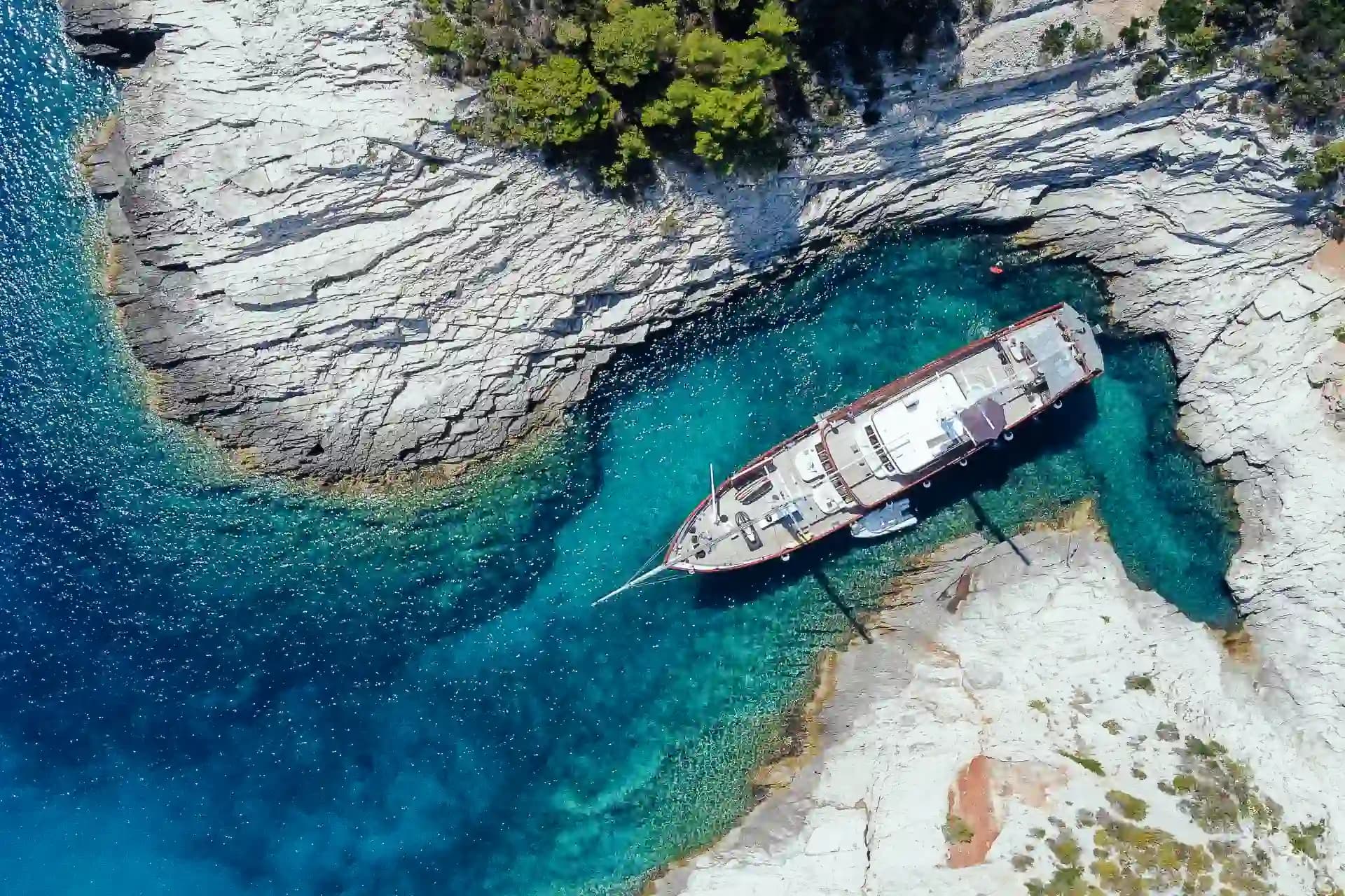 Aerial view of a large boat anchored in turquoise cove surrounded by white rocky coastline and green trees.