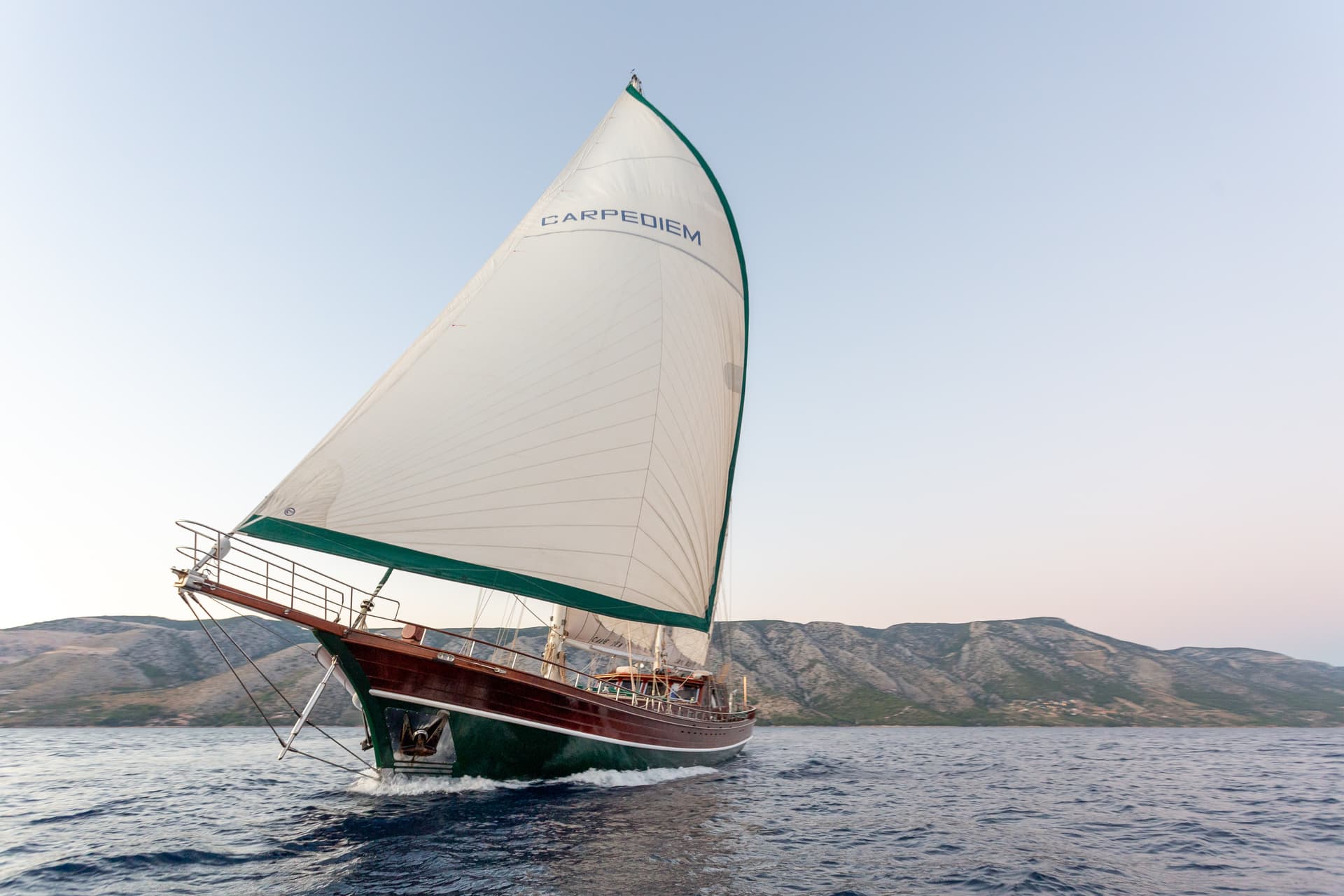 Wooden sailboat named CARPEDIEM sailing on dark blue water near arid coastline.