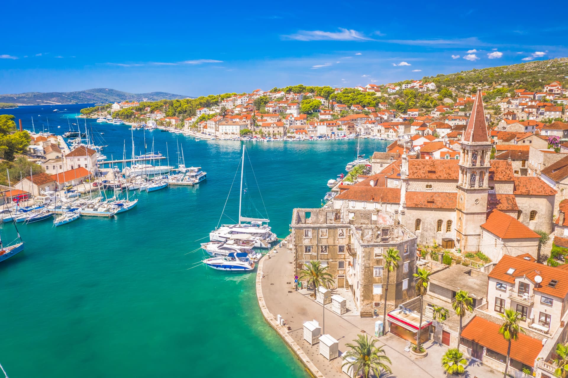 A drone view of Milna on Brač Island, Croatia, featuring the Church of Our Lady of Annunciation and its bell tower, a scenic bay filled with boats, a vibrant waterfront, and traditional Mediterranean